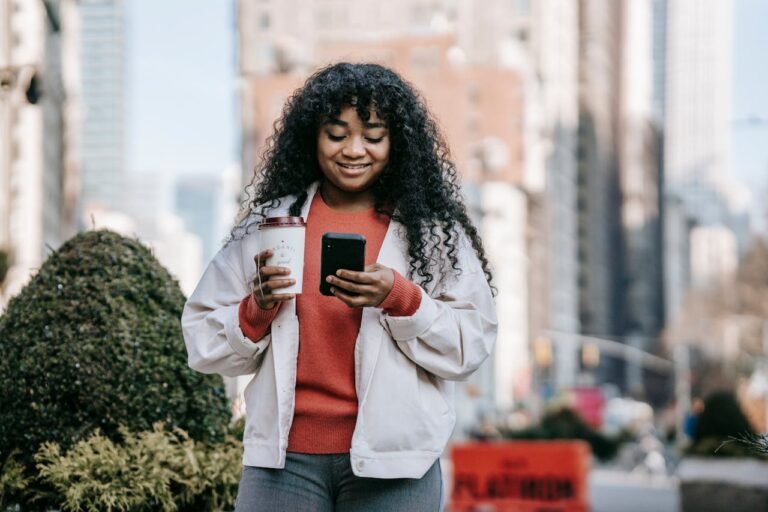 Femme noire souriante avec un café à emporter et naviguant sur son smartphone dans la rue